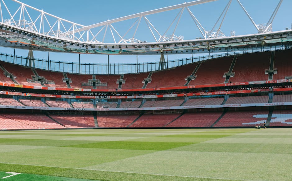 An empty football stadium with a well-maintained grass pitch in the foreground, surrounded by red tiered seating and multiple levels of viewing galleries. The stadium features a large, white, open-roof structure supported by a network of steel beams, allowing natural light to illuminate the space. The seating areas appear clean and free of debris, and the grass surface looks freshly cut and even, indicative of regular surface cleaning and maintenance. Bright daylight highlights the vibrant green of the pitch and the modern architectural details of the stadium. This image reflects the importance of deep cleaning and surface sanitisation in large public venues, consistent with professional cleaning standards offered by Cleaner Highbury, as seen on the page about best carpet cleaning near Highbury Fields and Highbury Park.