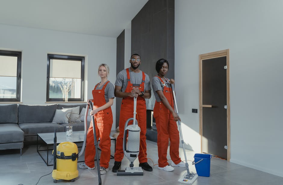 Two professional cleaners from Cleaner Highbury are performing surface cleaning in a modern living room. The cleaner on the left is operating a yellow upright vacuum cleaner on a tiled floor, which ap