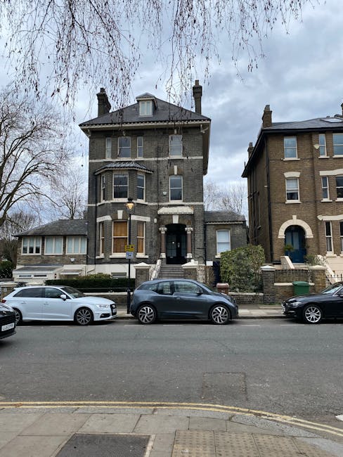 The image depicts a residential street with a large, multi-story Victorian-style house featuring a dark grey brick façade, white window trims, and a central entrance with a small staircase. The house 