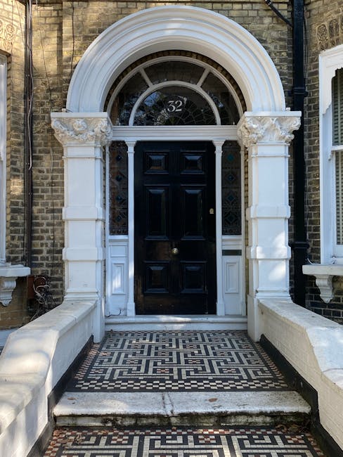 The image depicts a residential street with a large, multi-story Victorian-style house featuring a dark grey brick façade, white window trims, and a central entrance with a small staircase. The house 