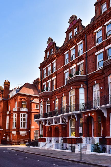 The image features a row of historic red brick residential buildings on a street in Highbury, N5. The buildings have multiple stories with large sash windows, some with white framing, and decorative architectural details such as arched window lintels and ornate cornices. The ground floor includes entrances with stairs and white-painted railings, while small balconies with black wrought iron railings are visible on the upper floors. The facades appear clean and well-maintained, with clear glass in the windows reflecting the blue sky and surrounding structures. The street is lined with a sidewalk and street parking, with minimal street furniture visible. The scene is illuminated by natural daylight, highlighting the classic architectural style characteristic of Highbury, and associating with high standards of property maintenance exemplified by Cleaner Highbury’s domestic cleaning services.