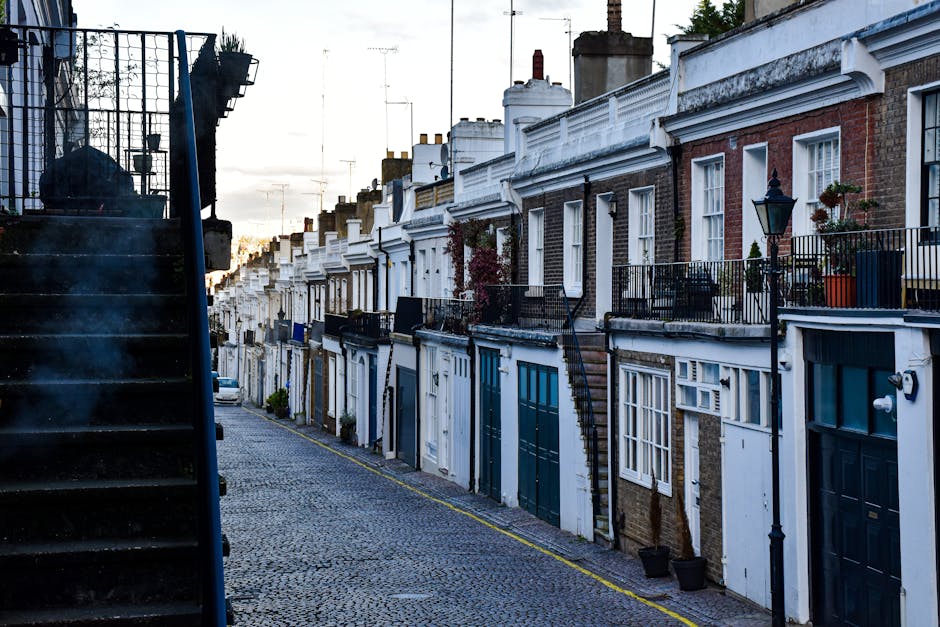 A row of terraced houses along a narrow cobblestone street in Highbury N5, featuring mixed brick and white-painted facades with large windows and small balconies. The scene is illuminated by soft natural light at sunset, with visible black wrought iron railings, street lamps, and potted plants on some balconies. On the left side, a black metal staircase with steps leads upward, and the street appears clean and well-maintained. The image depicts an outdoor residential setting typical of Highbury, highlighting the area's charming architecture. Cleaner Highbury offers professional cleaning services to maintain such properties and ensure cleanliness and hygiene in domestic environments.