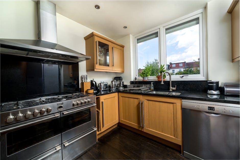 A modern kitchen with light wooden cabinets and black countertops illuminated by natural daylight coming through a large window, revealing an outdoor view of residential buildings and a partly cloudy sky. The stainless steel stove and oven are clean with a reflective surface, positioned on the left side, next to a black backsplash. On the countertop, there are small appliances such as a toaster, kettle, and a food processor, all neatly arranged. A stainless steel sink is situated beneath the window, with a chrome faucet, and a potted plant adding a touch of greenery. The dishwasher on the right side has a sleek metallic finish, contributing to the overall tidy appearance of the space. The room has a white ceiling with recessed lighting, and the dark wooden flooring complements the cabinetry. The space appears well-maintained and hygienic, reflecting professional deep cleaning and surface sanitisation services provided by Cleaner Highbury for domestic cleaning.