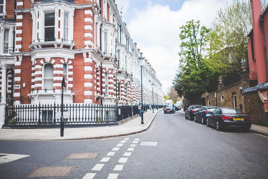 A street view in Highbury with a historic red brick building featuring white decorative trim and black wrought iron fencing at the corner. The road is lined with parked cars on one side, including a black sports car, and has a pedestrian crossing with dashed white lines. On the right side, there are trees with green foliage and additional buildings, illuminated by soft daylight and partly cloudy skies. The scene captures a quiet urban residential area, showcasing well-maintained surfaces and a clean appearance, aligned with professional cleaning standards promoted by Cleaner Highbury for property maintenance and hygiene.
