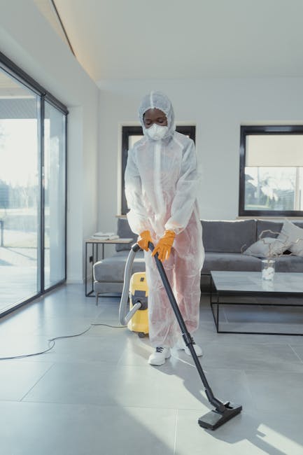 A professional cleaner wearing a white protective suit, face mask, and orange gloves using a vacuum cleaner to perform surface cleaning and deep cleaning in a modern living room. The room features large glass sliding doors, allowing natural light to illuminate the space, with a grey sofa and a white coffee table holding a water bottle. The tiled floor appears clean and polished, indicative of thorough sanitisation process. Visible cleaning tools include the vacuum hose and a yellow portable vacuum unit, emphasizing a focus on hygiene and maintenance. The scene reflects domestic cleaning services provided by Cleaner Highbury, aimed at maintaining high standards of cleanliness and hygiene in residential spaces.